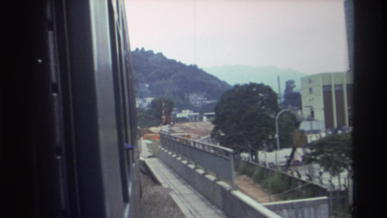 View from a moving train window showing a concrete trackside barrier, green hills, hillside buildings, trees, and a tall building at the edge.