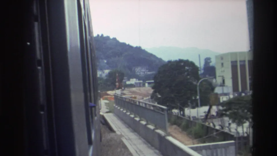8mm train window footage from Hong Kong, 1982, showing a concrete lineside barrier, terraced green hills, scattered hillside buildings, and a tall modern building at the frame edge.