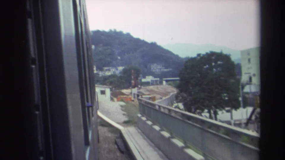 8mm train window footage from Hong Kong, 1982, showing trackside construction earthworks, a small white building, a large deciduous tree, and hazy hillside housing receding into the background.