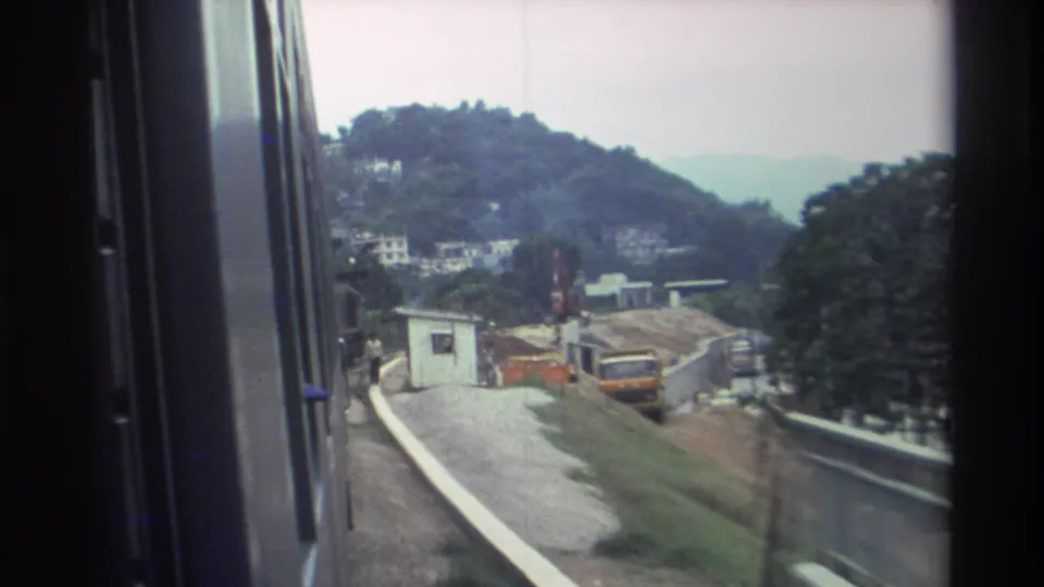 8mm footage from a moving train in Hong Kong, 1982, showing a yellow excavator or construction vehicle at a trackside site, with terraced hills and hillside buildings beyond.