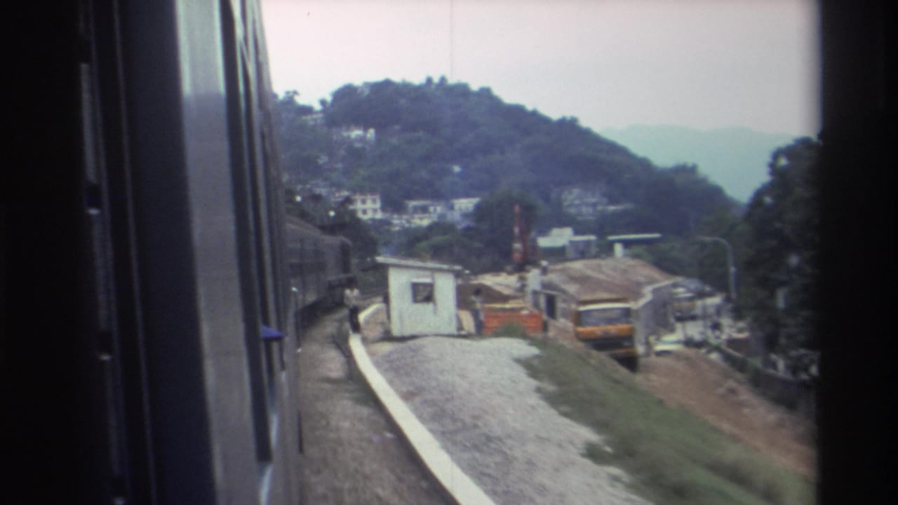 8mm train window view, likely 1982 Hong Kong: small white station kiosk beside a construction site on a wooded hillside, orange barriers and yellow vehicle visible.