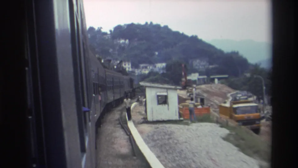8mm train window POV of a small white kiosk and yellow construction truck beside the rail line on a Hong Kong hillside, approximately 1982.