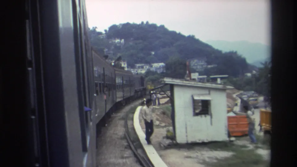 8mm train window view of a worker walking trackside near the construction kiosk and orange barriers on a Hong Kong hillside, c. 1982.