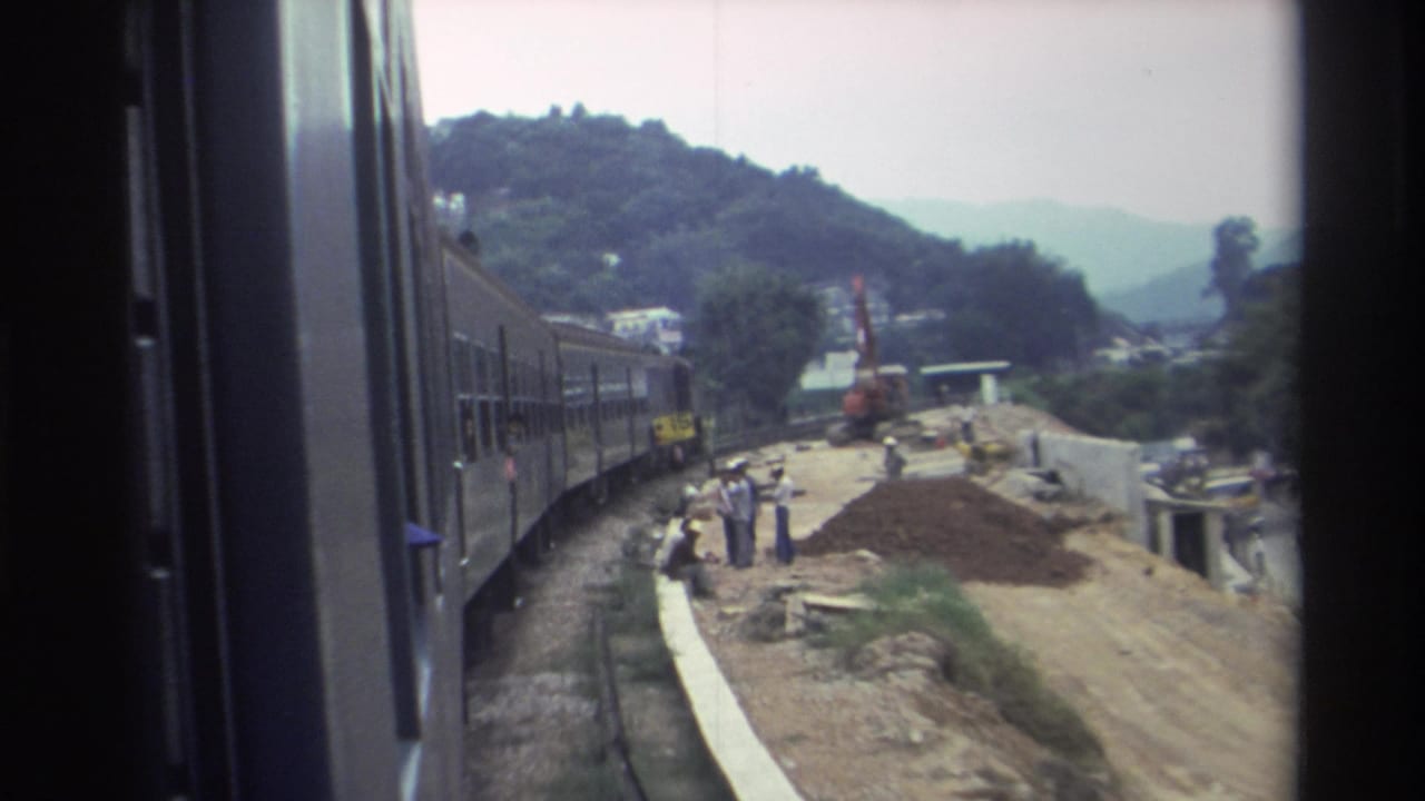 1982 Hong Kong — train-door view of a curved rail line passing a construction site with reddish dirt mounds, workers, and an orange crane against forested hills.