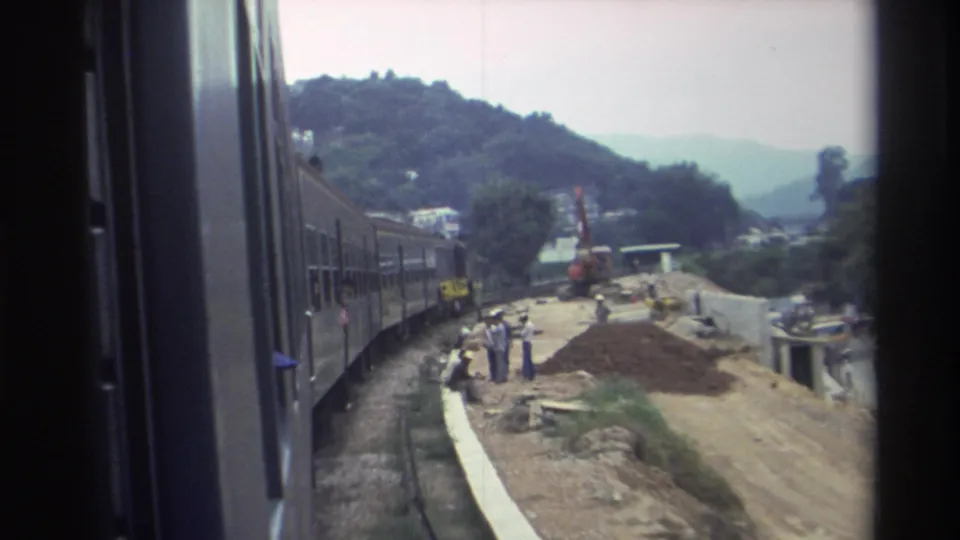 1982 Hong Kong — train-door view of a curved rail line passing a construction site with reddish dirt mounds, workers, and an orange crane against forested hills.