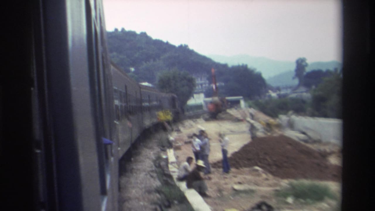 Workers in light-coloured clothing stand near reddish dirt mounds at a rail-adjacent construction site, viewed from a passing train, with hills and a red vertical element behind.