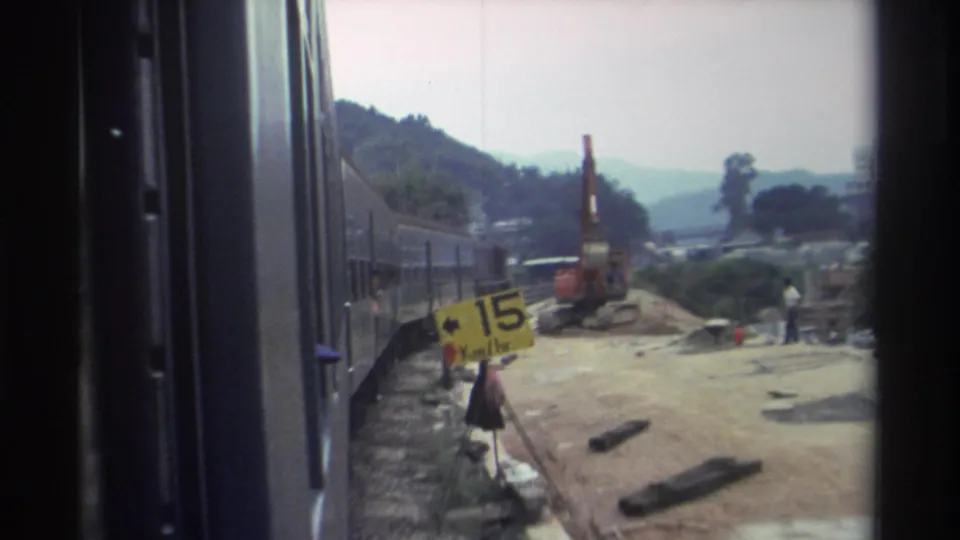 1982 Hong Kong — yellow '15 Km/hr' speed-restriction sign beside the rail track, with orange construction machinery and a worker visible in the background.