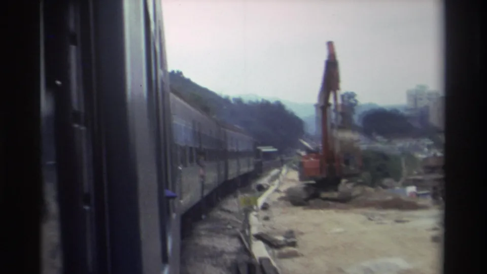 8mm footage, c.1982, likely Hong Kong — long train curving past earthworks with an orange excavator and apartment buildings in the background.