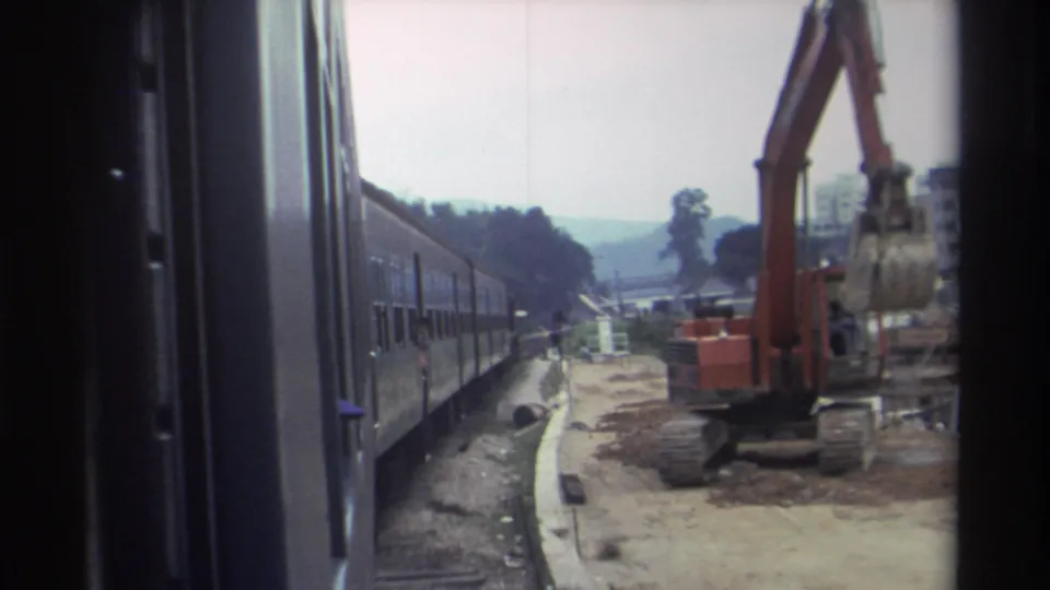 8mm footage, c.1982, likely Hong Kong — large orange hydraulic excavator operating alongside the railway, workers and construction materials on bare ground.