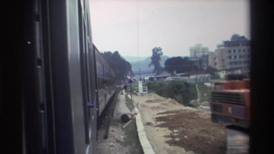 8mm footage, c.1982, likely Hong Kong — red construction vehicle alongside the railway; residential apartment blocks and trees in the background.