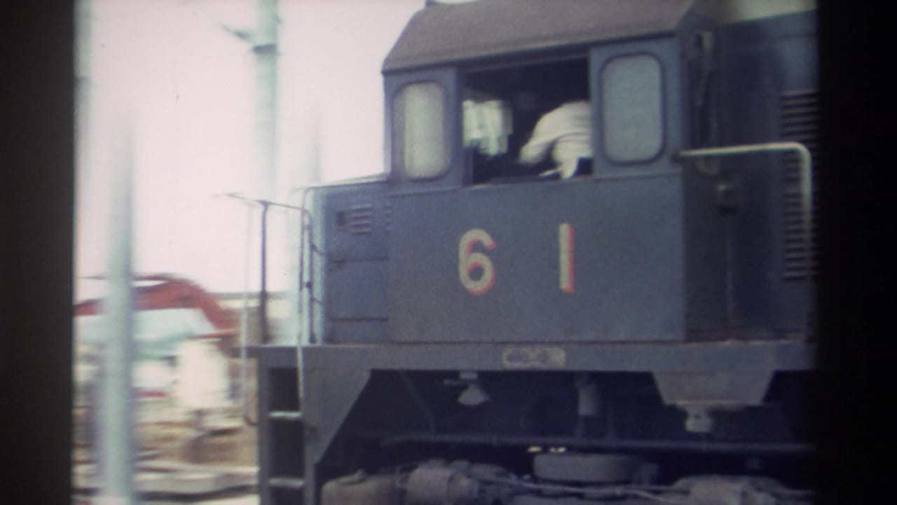 Locomotive 61 in motion, figure visible in cab window, construction equipment in background.