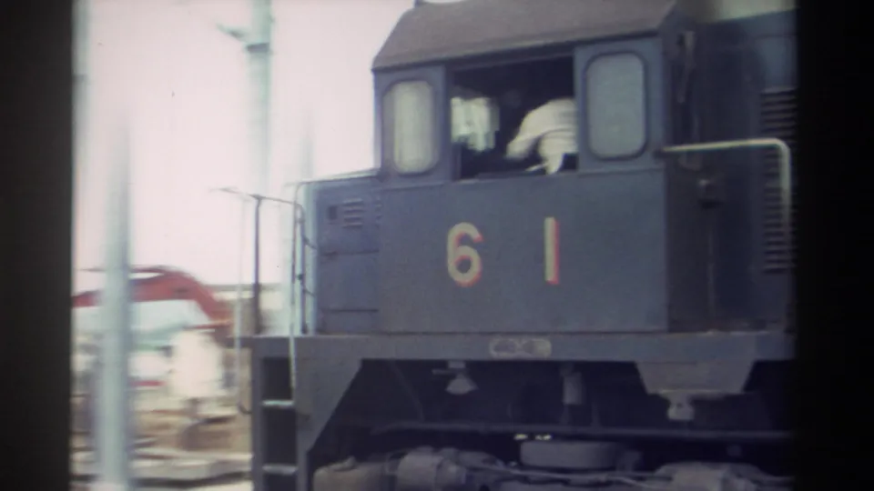 Blue-grey diesel locomotive 61 moving, figure in cab window, construction equipment and utility poles visible behind, 1982 Taiwan.