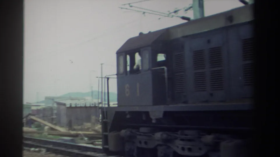 Diesel locomotive 61 moving away along railway yard tracks, green overhead catenary poles and industrial skyline visible, 1982 Taiwan, 8mm.