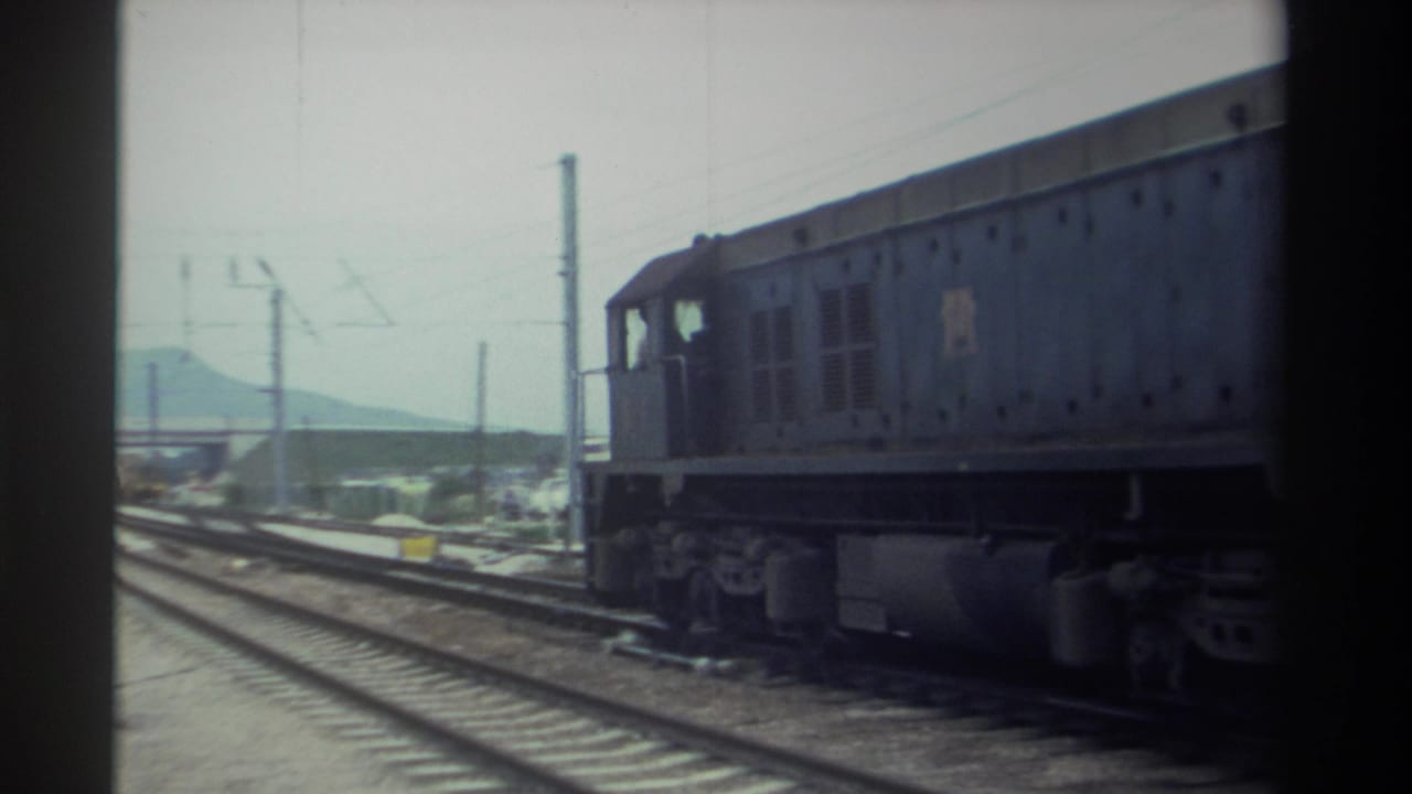 Dark blue diesel locomotive on multi-track railway with overhead catenary wires and hazy mountains in the background.
