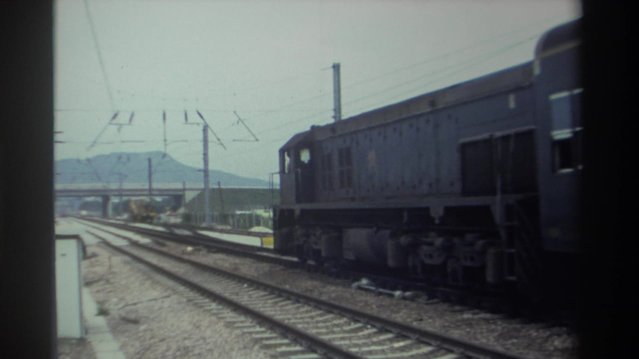 Blue-grey diesel locomotive on multi-track rail yard, overhead catenary wires, mountain ridge visible through haze.