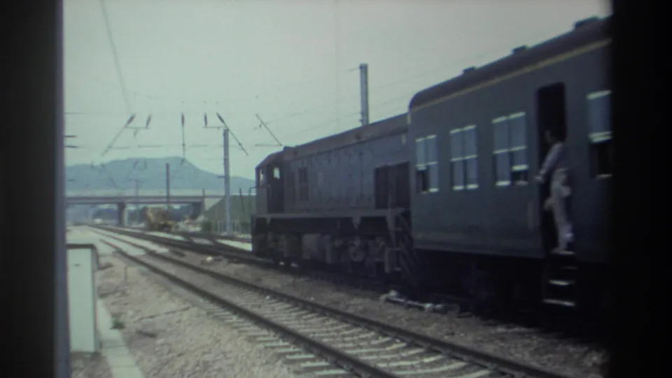 8mm home movie frame, c.1982 Taiwan — passenger train with a figure visible at an open carriage door, overhead catenary wires, distant mountains.
