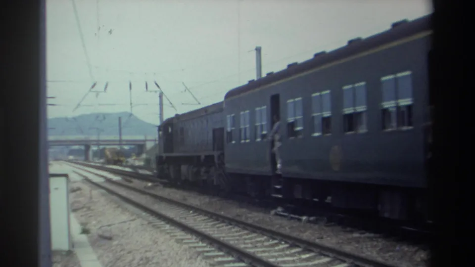 8mm home movie frame, c.1982 Taiwan — passenger train cars with a figure at an open door, overhead wires, and a hazy mountain silhouette in the background.