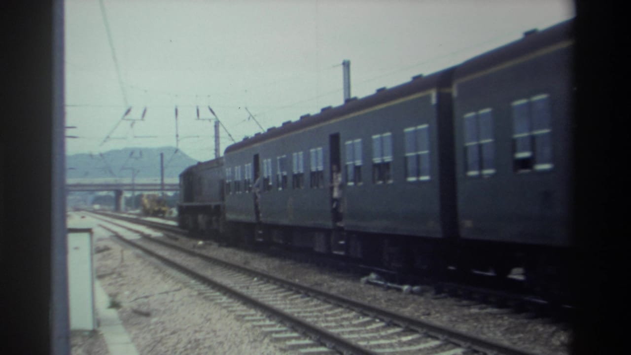 Taiwan, 1982 — passenger train with electrified overhead lines; figure near open doorway; low hills and what appears to be a viaduct or bridge in background.