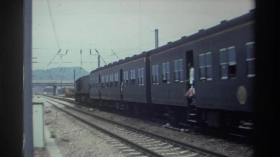 Taiwan, 1982 — multi-car passenger train on electrified railway receding into distance; figure near open doorway; overcast sky and low hazy hills.
