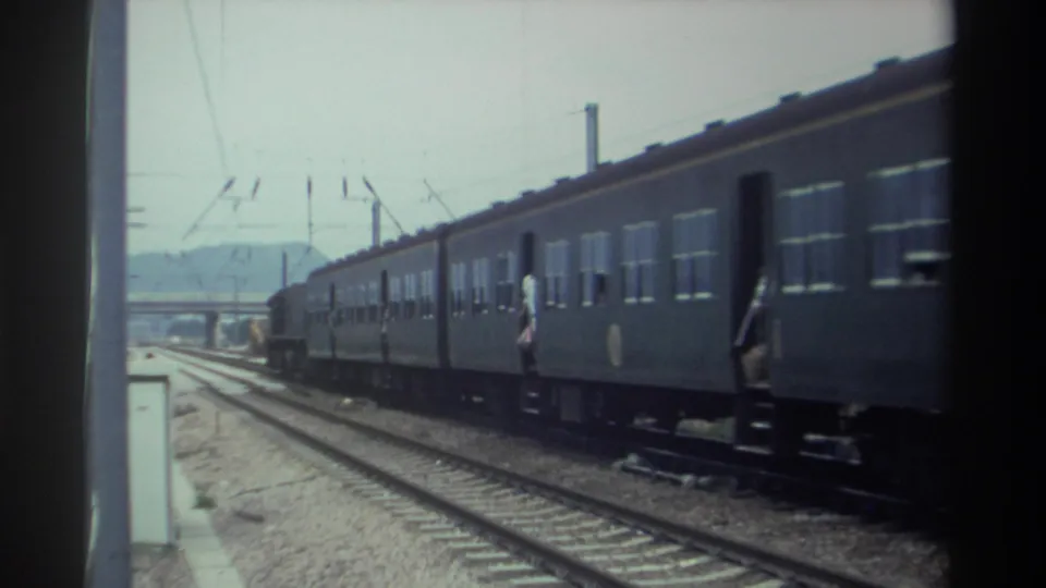 Taiwan, 1982 — passenger train on electrified multi-track line continuing past; figure near open coach doorway; low hills and overcast sky.
