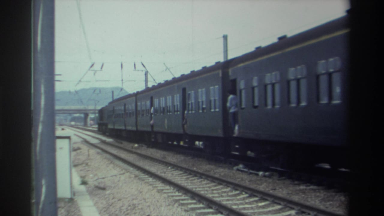 Dark green passenger train with figure standing near open door, catenary poles and wires overhead, distant hills visible, Taiwan 1982, 8mm.