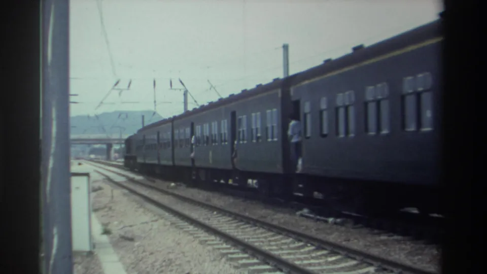 Dark green passenger train with figure standing near open door, catenary poles and wires overhead, distant hills visible, Taiwan 1982, 8mm.