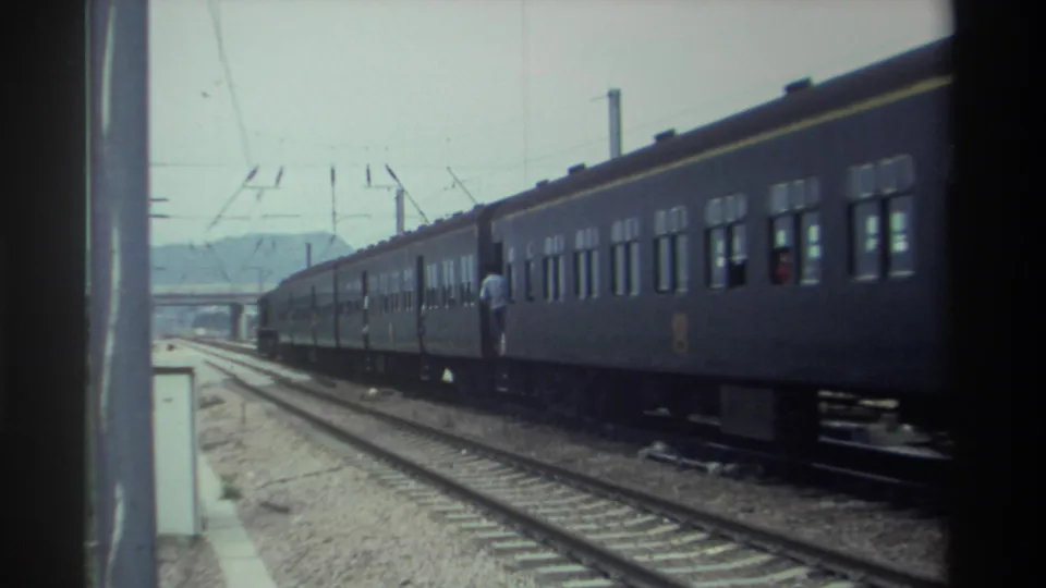 Dark green passenger train with yellow trim stripe and a figure in light clothing standing beside it, overhead catenary wires, Taiwan 1982, 8mm.