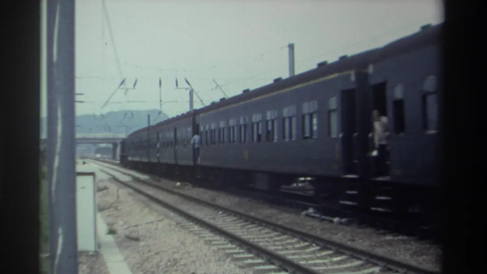 Dark green passenger train with multiple figures near doorways, overhead electric wires, bridge structure and hills in background, Taiwan 1982, 8mm footage.