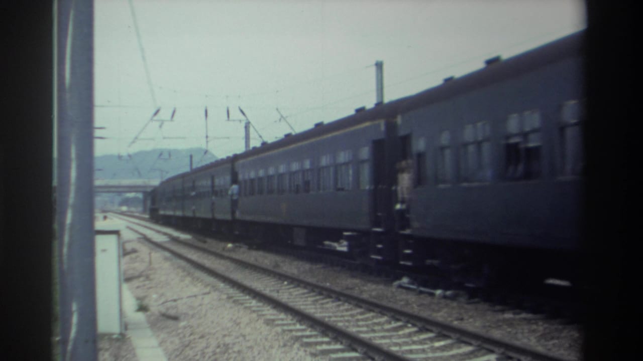 Dark gray passenger train moving along electrified railway, shot from platform level, with overhead catenary wires visible.
