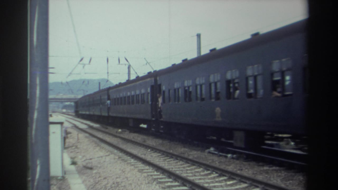 Gray passenger train rolling past a station platform with figures visible alongside, Taiwan, 1982; catenary poles and distant hazy hills in background.