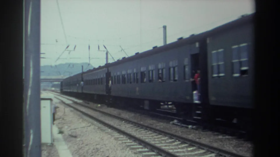 Gray passenger train with person in red on car steps and a figure alongside the track, Taiwan, 1982; 8mm film grain and edge scratches visible.