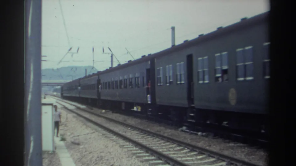 Rear carriages of gray passenger train passing with a figure visible trackside, Taiwan, 1982; 8mm film scratches and grain on left edge.