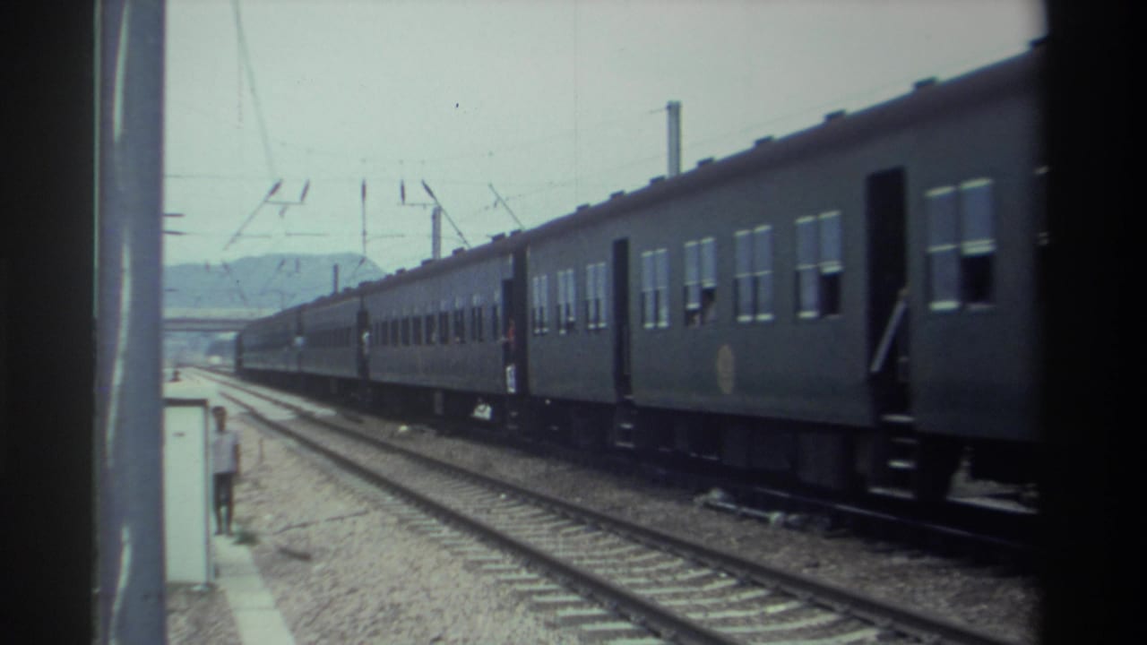 8mm home movie frame from Taiwan, 1982, showing a long dark green passenger train passing a platform with overhead catenary electrification and a figure standing near the edge.