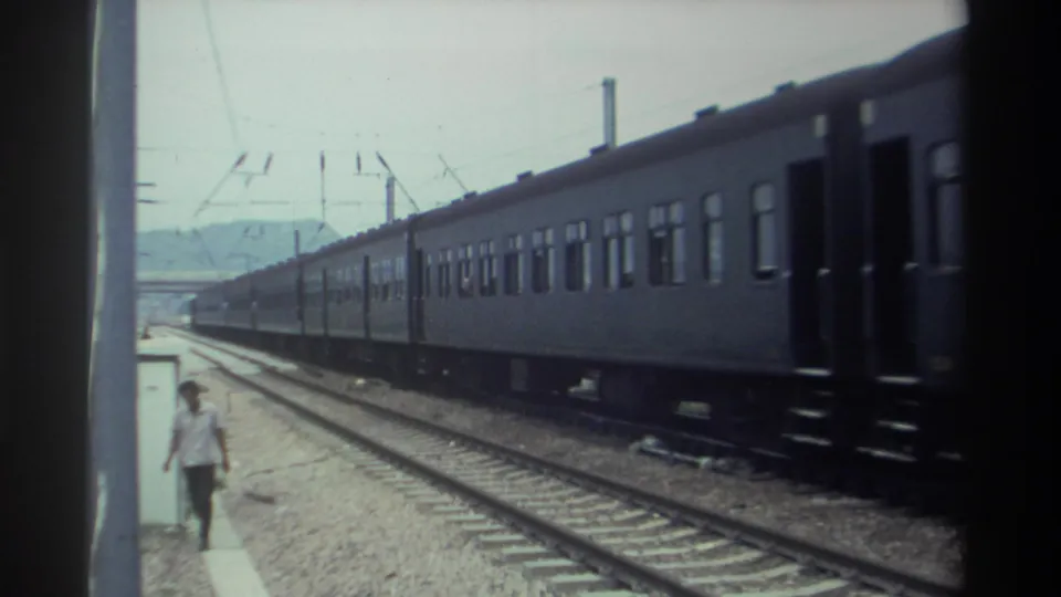 8mm footage from Taiwan, 1982, showing a man walking along a station platform as a long electrified passenger train passes alongside, catenary wires overhead and hills faintly visible in the distance.