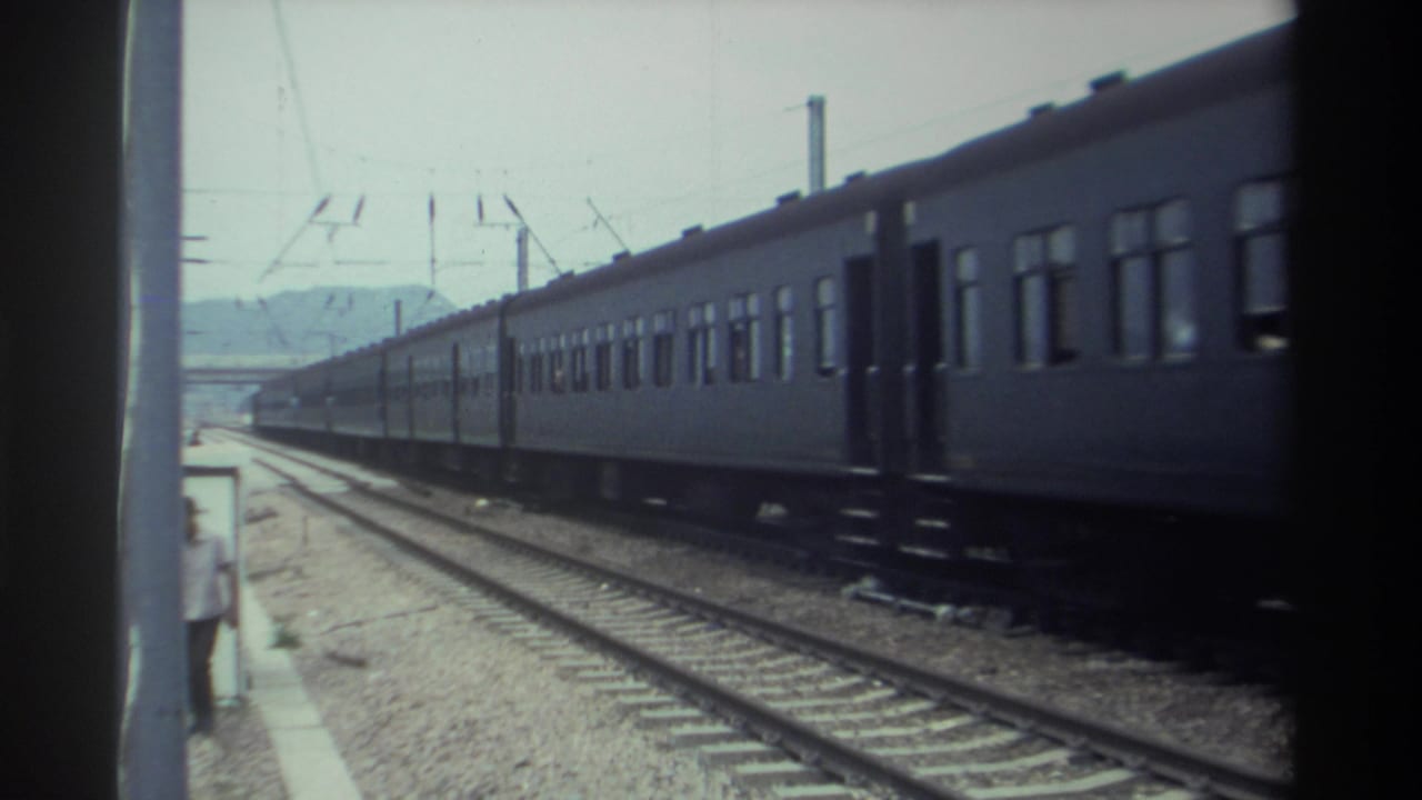 1982 Taiwan — dark blue passenger train moves past a gravel-ballast platform, overhead catenary wires visible, hazy hills in the distance, shot on 8mm film.