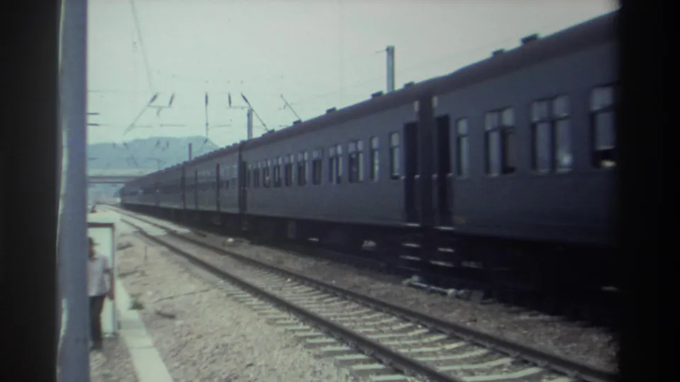 1982 Taiwan — dark blue passenger train moves past a gravel-ballast platform, overhead catenary wires visible, hazy hills in the distance, shot on 8mm film.