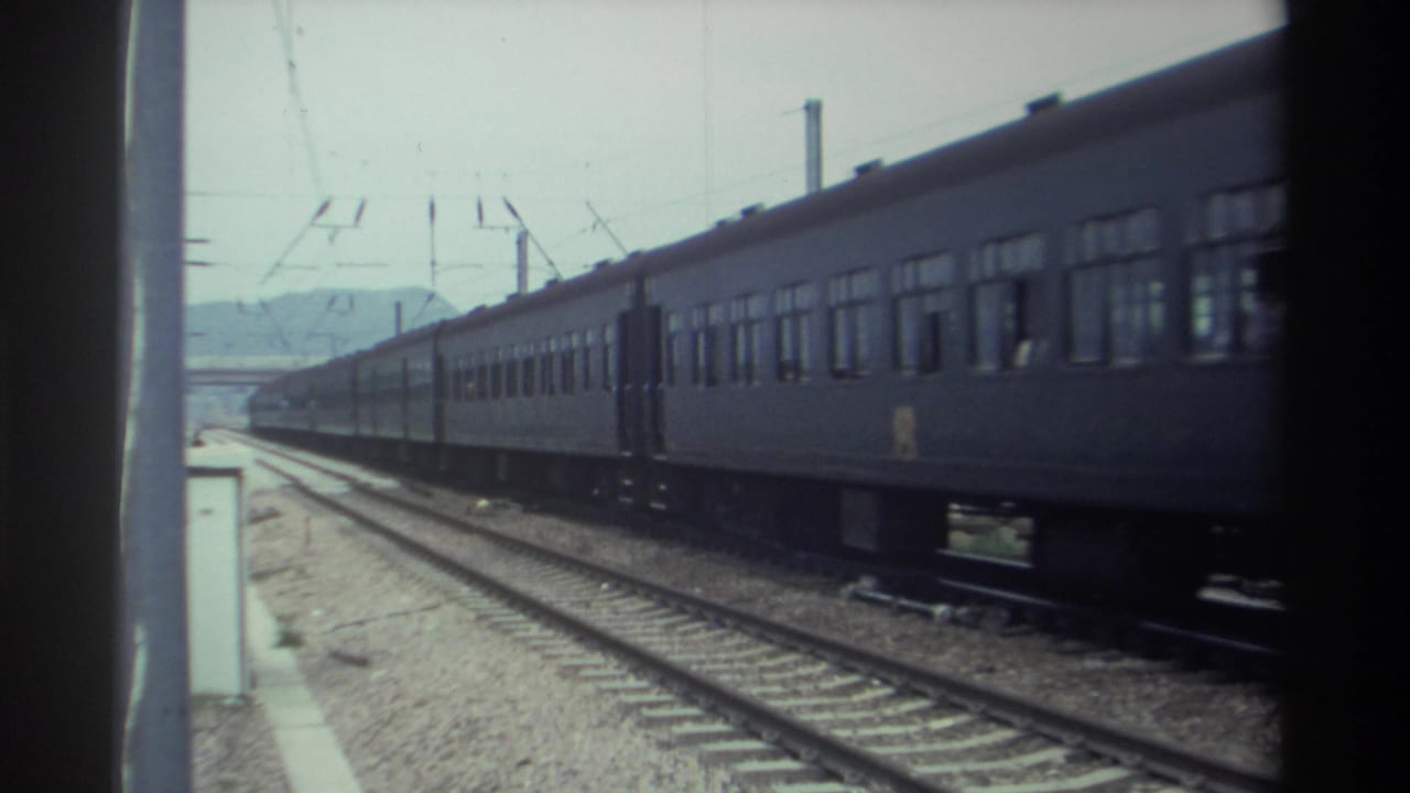 1982 Taiwan — blue passenger train carriages pass the platform edge, catenary wires overhead, hazy hills behind, captured on 8mm film.