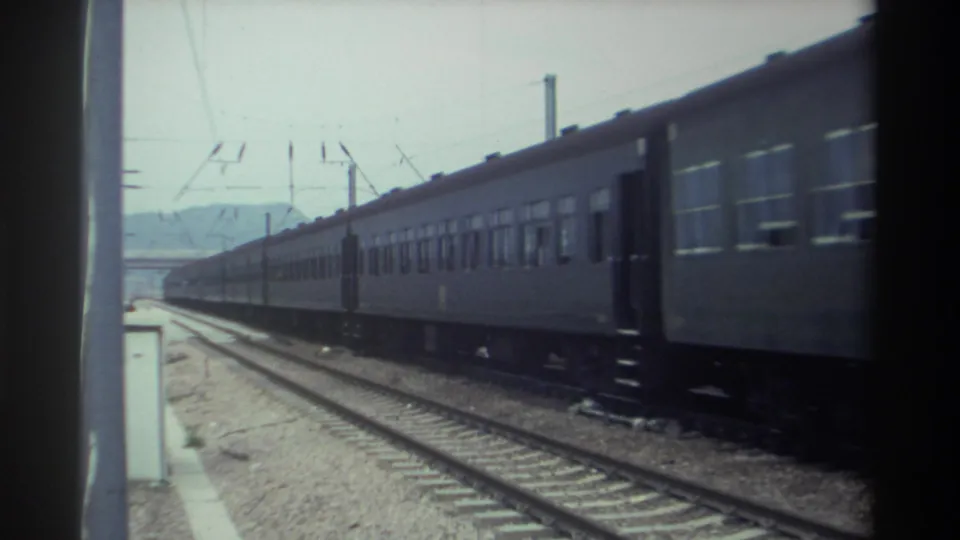 1982 Taiwan — long blue passenger train passes the gravel-side platform in mid-shot, catenary infrastructure overhead, hazy mountain horizon behind, on 8mm film.