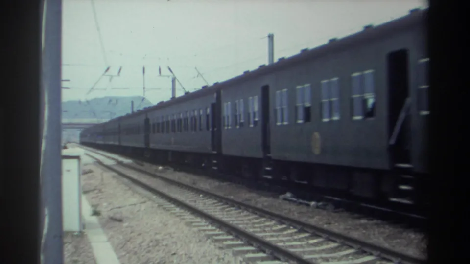 1982 Taiwan — final carriages of a blue passenger train pass on 8mm film, open parallel tracks and a hazy mountain horizon coming into view.