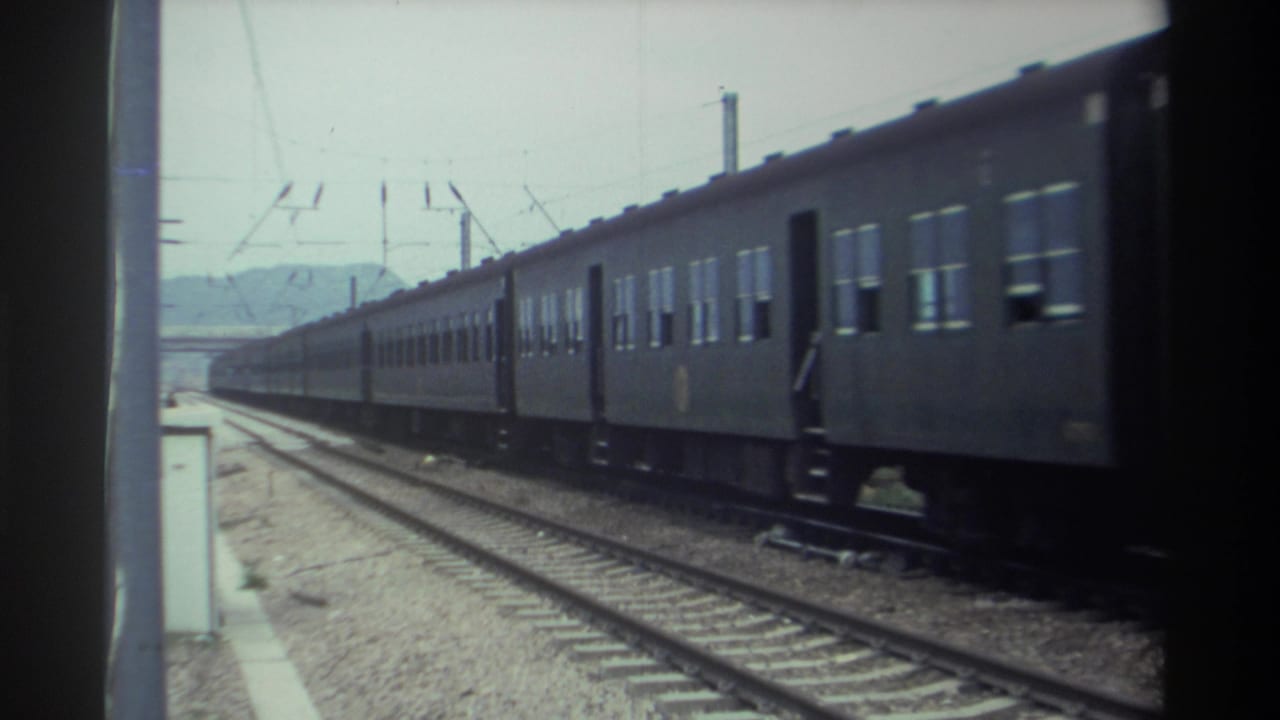 Long dark-painted passenger train passing a station platform in Taiwan, circa 1982, with overhead catenary visible, captured on 8mm film.