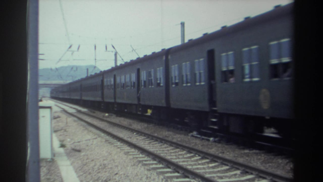 Passenger train carriages in motion past a station platform, overhead electrical catenary visible against an overcast sky.