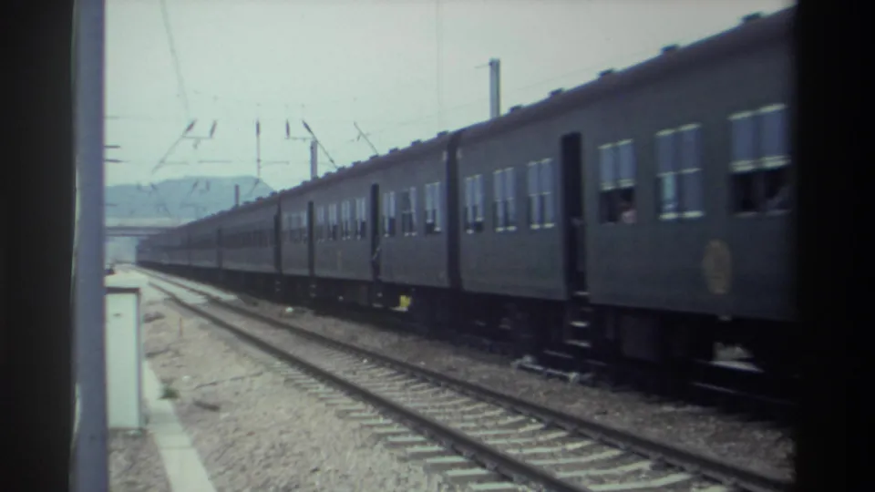Dark-painted passenger train carriages moving past a platform in Taiwan, circa 1982, showing catenary overhead system and distant hills, 8mm film.