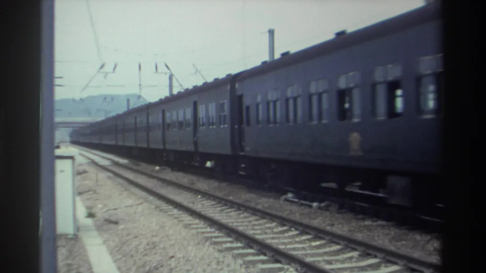 Rear cars of a dark-painted passenger train at a station in Taiwan, circa 1982, with hills and catenary visible, 8mm film.