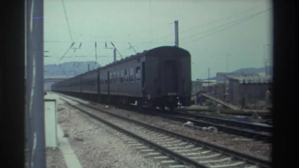 Tail end of a Taiwan passenger train departing frame, revealing open electrified track and a distant structure, circa 1982, shot on 8mm.