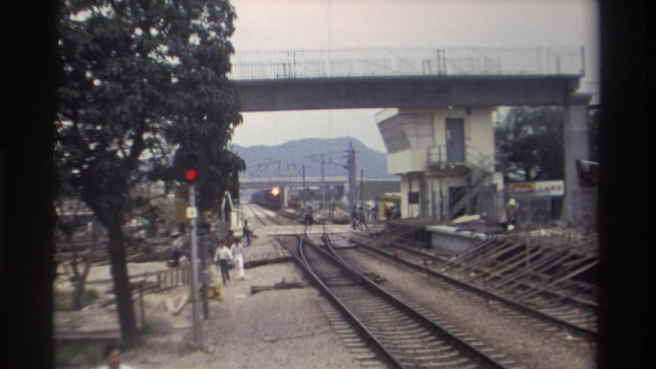 Track-level view of a Taiwan railway station circa 1982, showing multiple tracks, a red signal light, a concrete overpass, and pedestrians walking toward the platform.