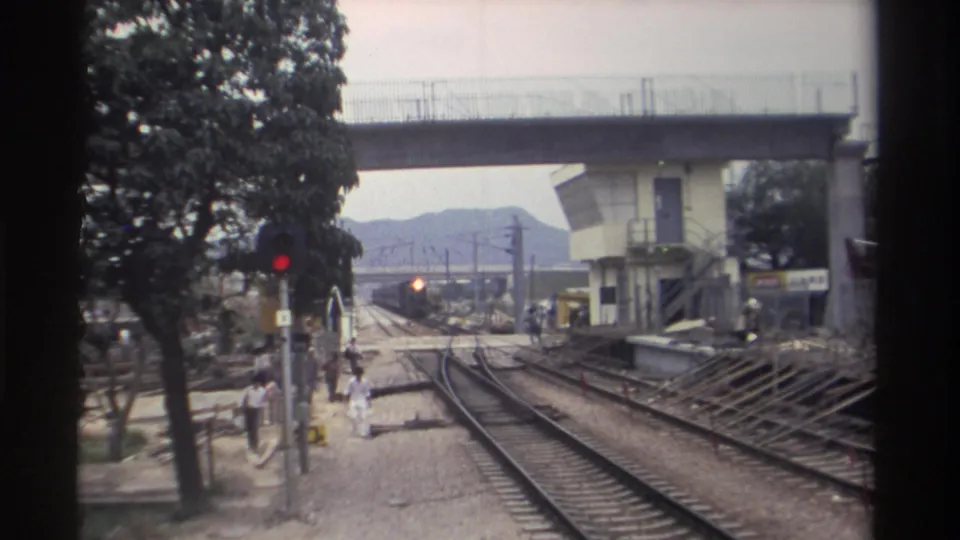 Taiwan railway station circa 1982; an approaching train becomes visible in the middle distance as pedestrians in casual dress walk alongside the tracks.