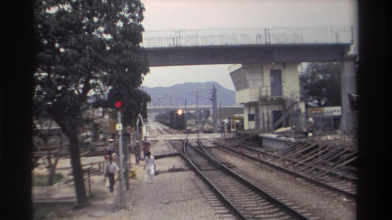 8mm film frame, Taiwan 1982: railway trackside looking toward an approaching train, with a concrete overpass above, red signal light, large trees, and pedestrians in casual clothing.