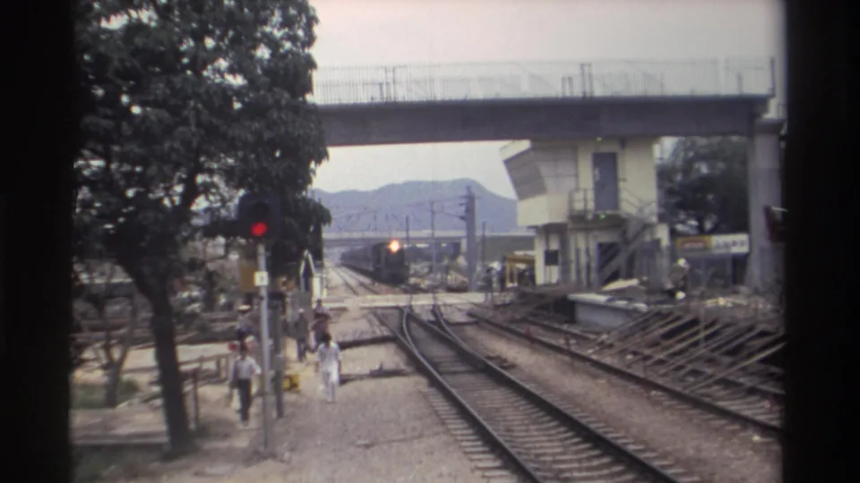 8mm film frame, Taiwan 1982: railway trackside looking toward an approaching train, with a concrete overpass above, red signal light, large trees, and pedestrians in casual clothing.