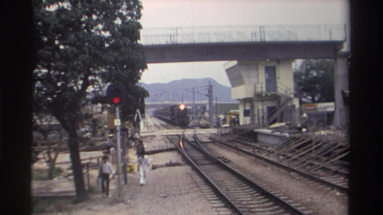 8mm footage, Taiwan 1982: railway trackside with train headlight growing brighter in distance, red signal light, concrete overpass, trees, and pedestrians on gravel path.