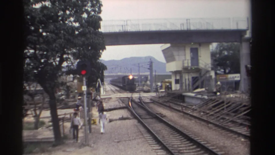 8mm footage, Taiwan 1982: railway trackside with train headlight growing brighter in distance, red signal light, concrete overpass, trees, and pedestrians on gravel path.